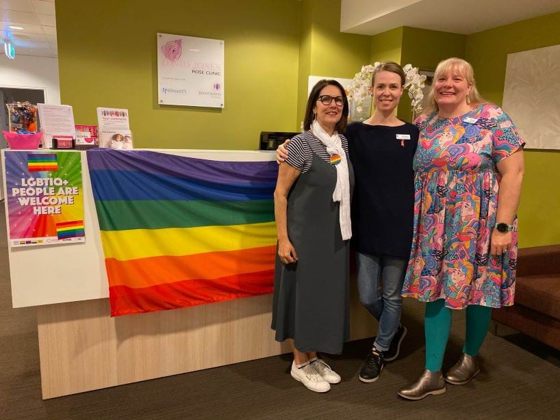 2019 Rainbow Rose screening session (provided by BreastScreen Victoria) Three people similing and standing in front of a rainbow flag with a sign saying LGBTIQ+ people are welcome here.
