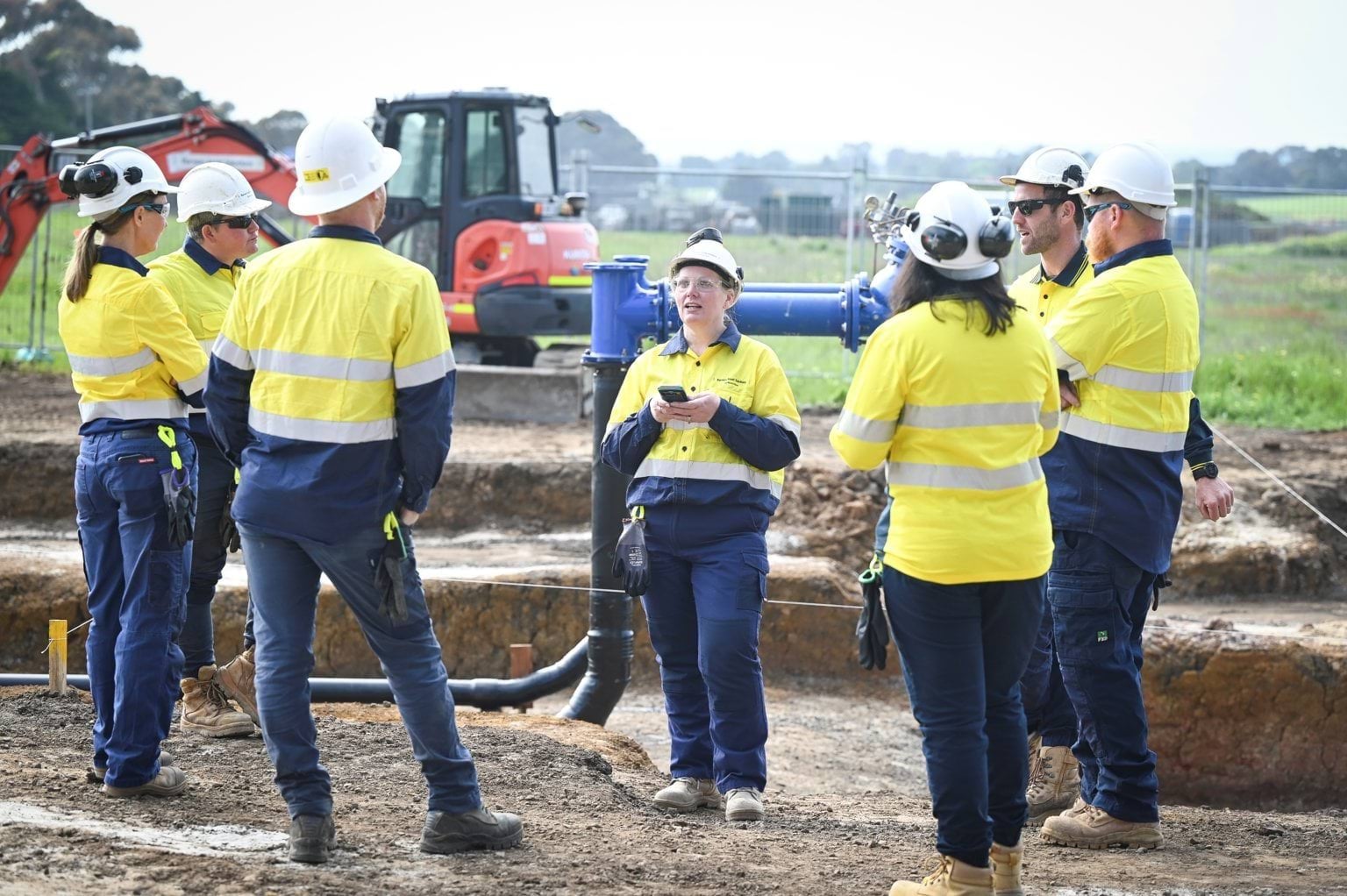 A group of construction workers, wearing yellow high-visibility safety gear and hard hats, stand in a circle having a discussion at an outdoor worksite with excavation equipment and pipes in the background.