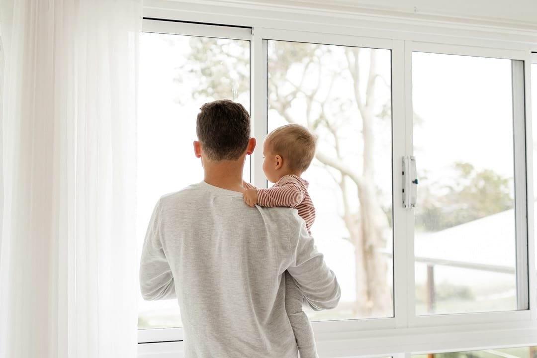 A father holding his young child while looking out a large window during the day. The child is resting their hand on the father's shoulder.