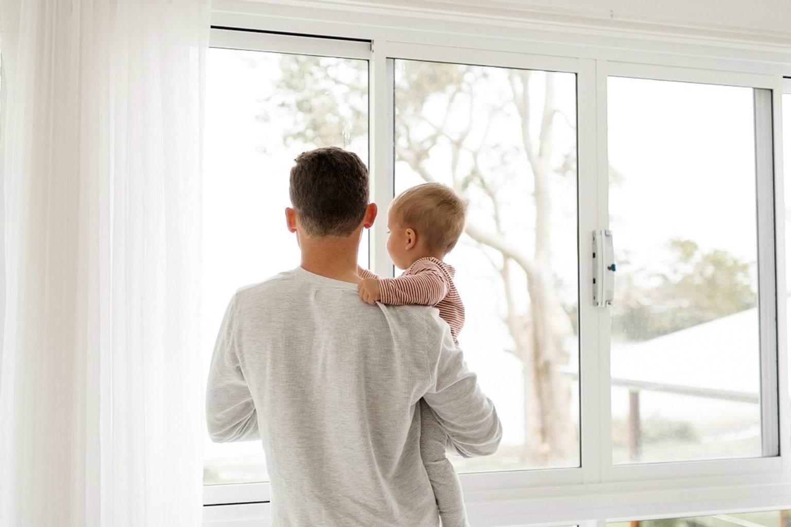 A father holding his young child while looking out a large window during the day. The child is resting their hand on the father's shoulder.