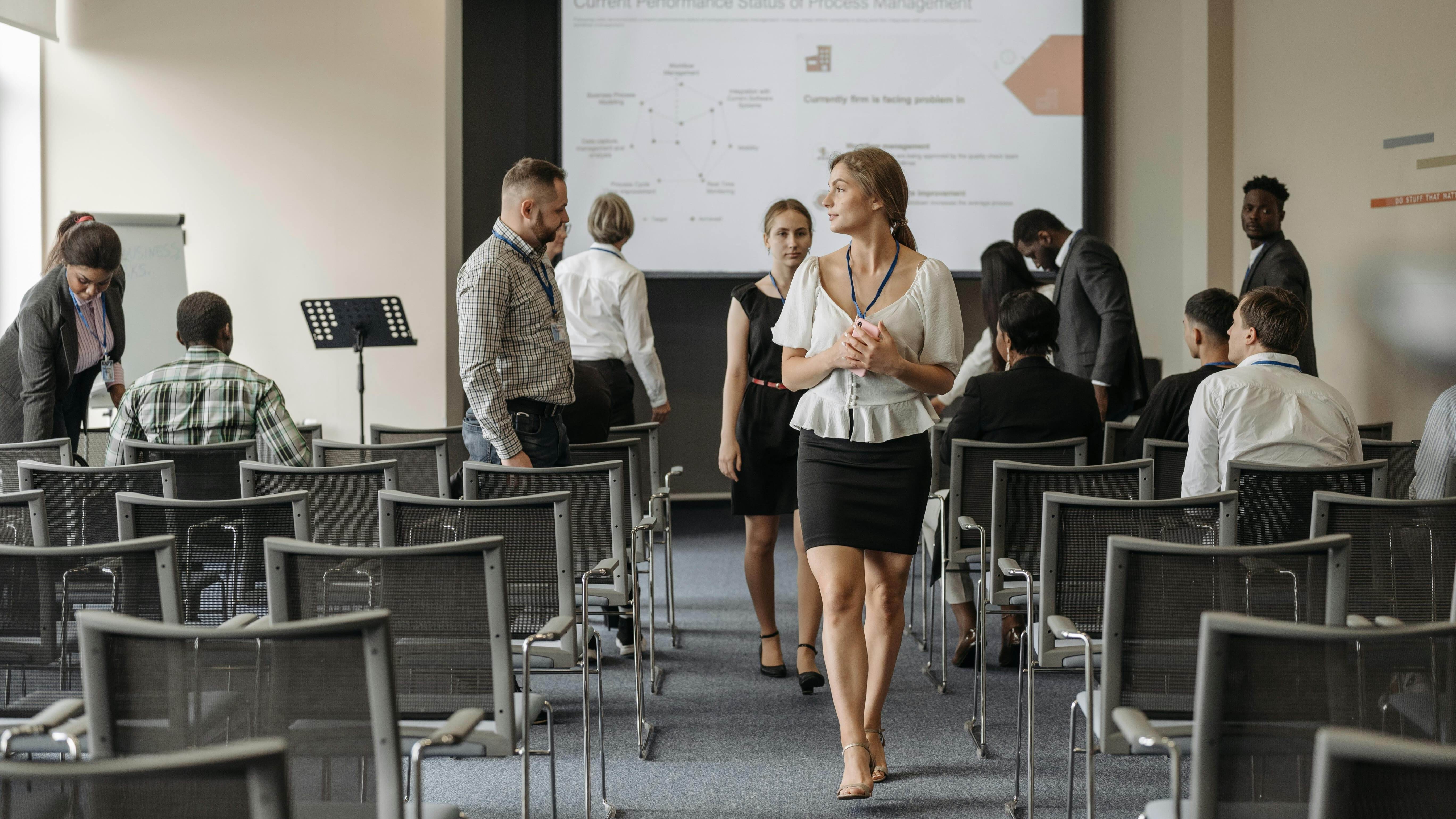People in business attire gather and talk in a conference room after a presentation, with chairs and a large screen in the background.