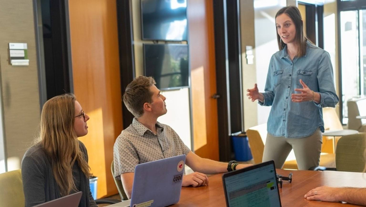 A group of people sitting at a meeting table with laptops. A woman is standing and speaking to the group.
