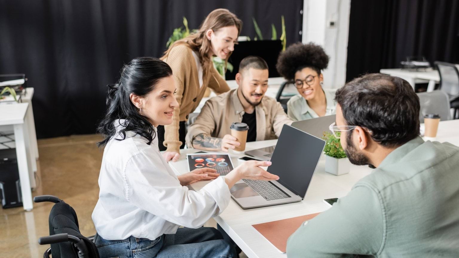cheerful team lead in wheelchair pointing at laptop, sharing ideas with man near business people